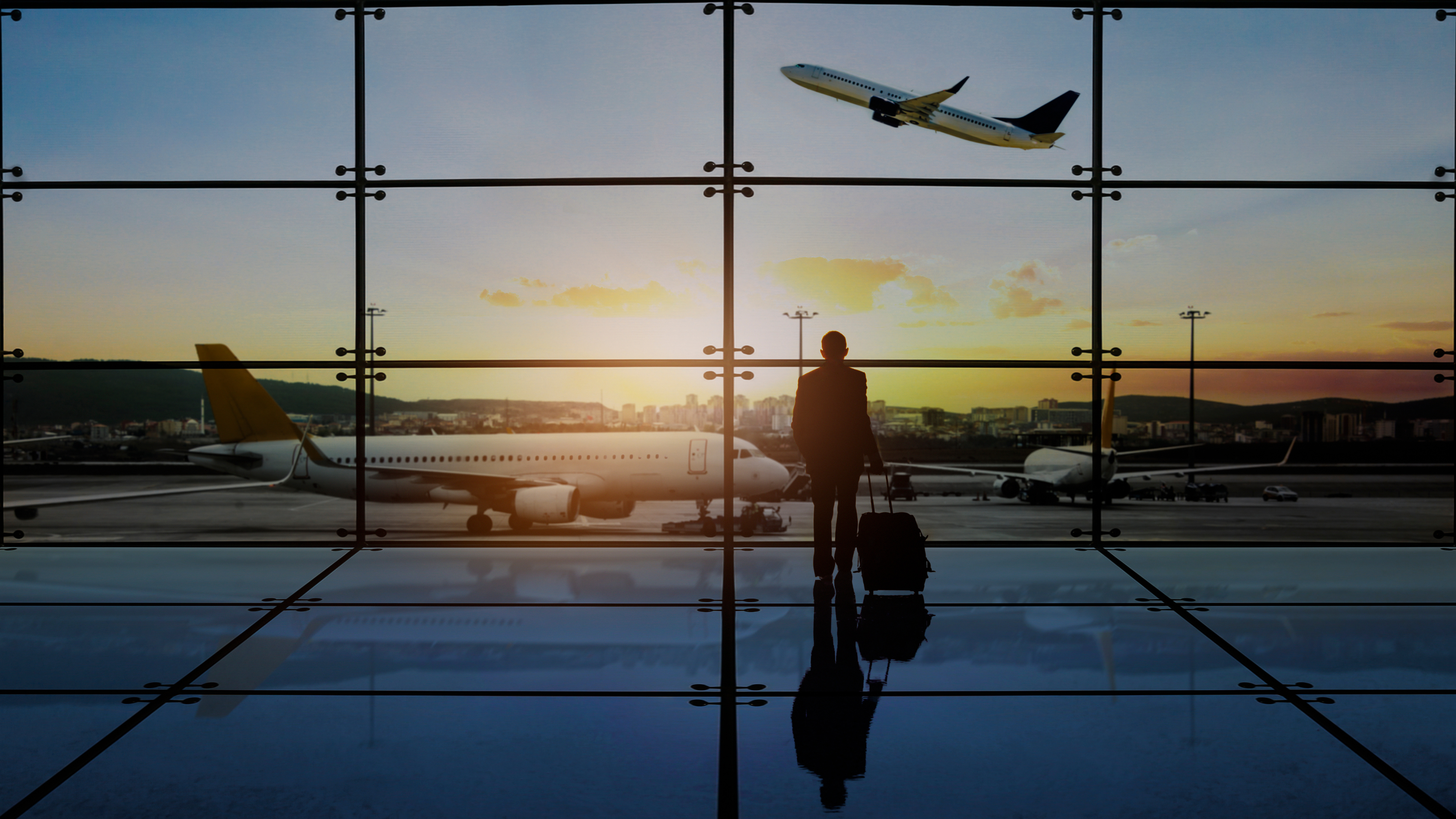 Business traveler with luggage silhouetted against floor-to-ceiling airport windows at sunset, aircraft on tarmac